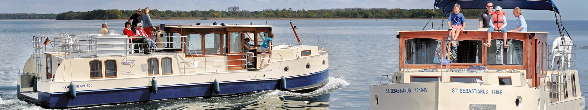 Zwei Hausboote auf denen jeweils eine Familie Urlaub macht fahren auf einem See aneinander vorbei. 